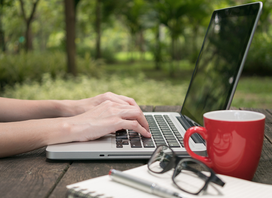 Blog - Close Up of Person Typing on a Laptop While Sitting Outside with a Red Mug, Note Book, and Glasses Sitting on the Wooden Table